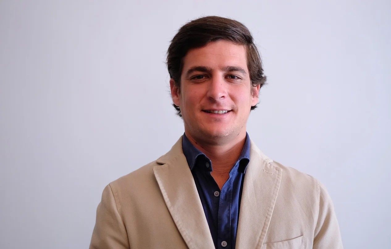Person wearing a beige blazer over a blue button-down shirt against a plain background.