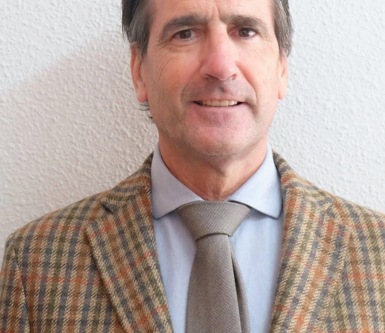 Man in plaid blazer and tie smiling against a textured wall.